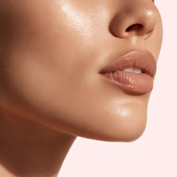 Close-up of a model's lower face and neck wearing a glossy, warm nude lipstick against a soft beige background.